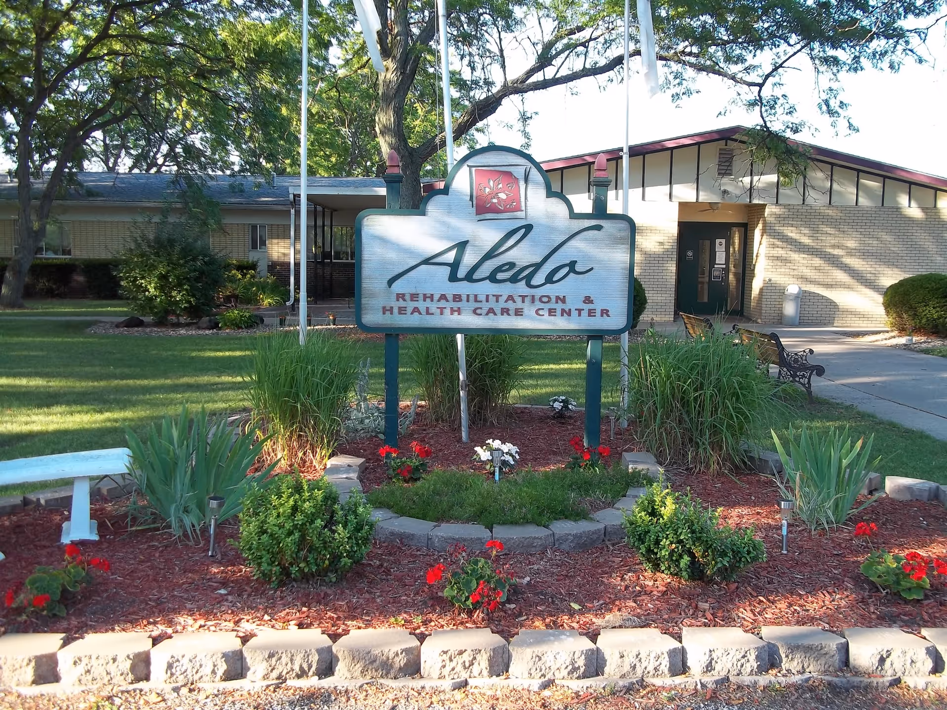 Outdoor view of the Aledo Rehabilitation & Health Care Center sign surrounded by landscaped plants and flowers, with a building and trees in the background.