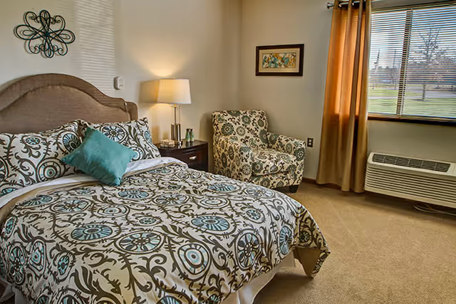 Well-lit bedroom with a patterned bedspread and upholstered headboard, nightstand and lamp, patterned armchair, wall art, and a window with curtains.