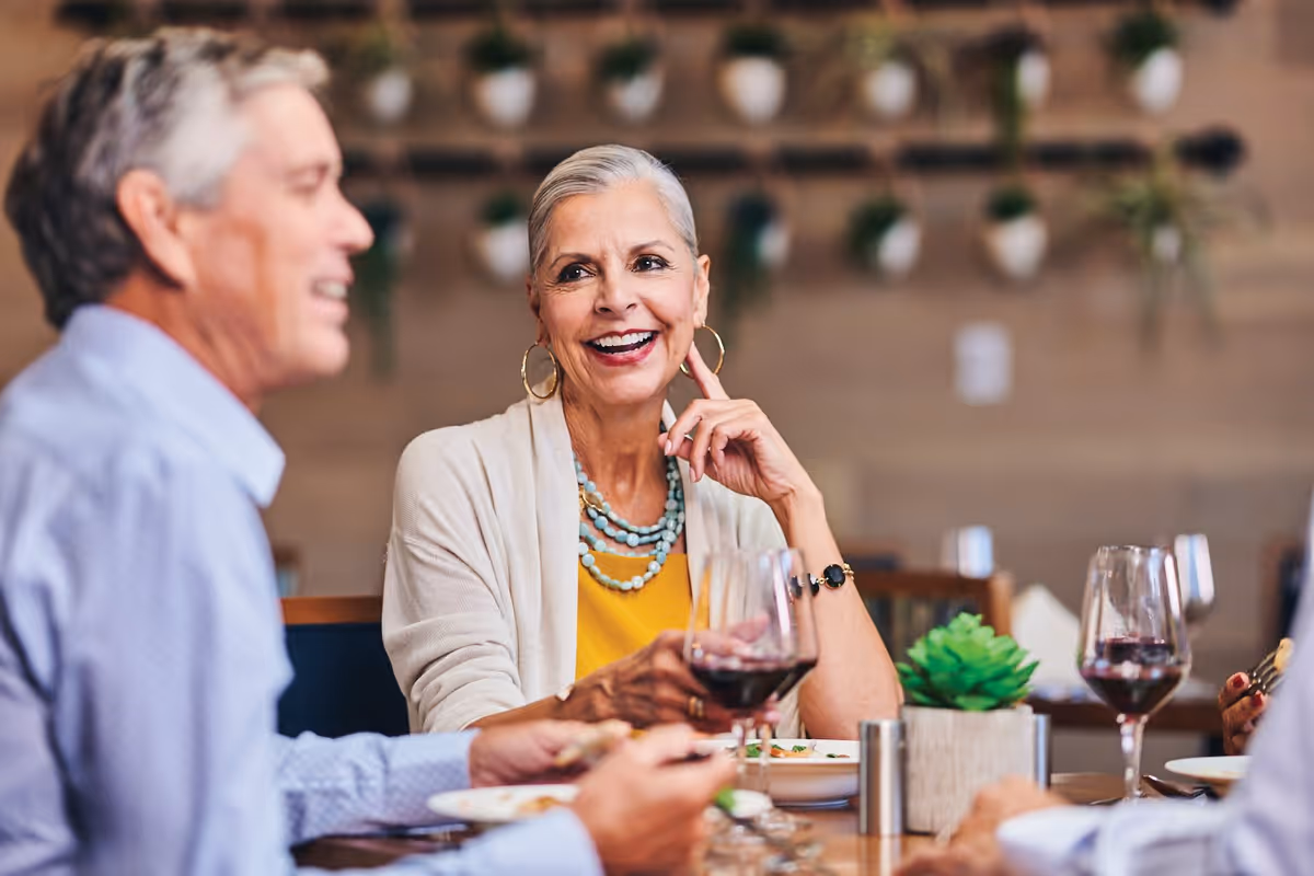Two elderly people sitting at a dining table, enjoying a meal and wine, with a woman smiling and engaging in conversation. The background shows a wall with hanging plants.
