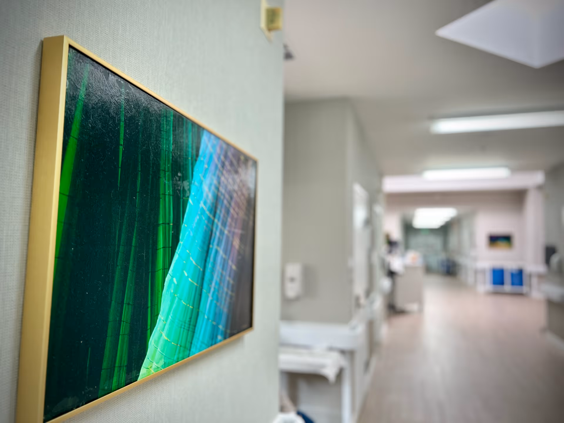 A framed abstract painting with green and blue hues hanging on a light-colored wall in a hallway of a senior living facility. The hallway is well-lit with fluorescent lights and has a clean, modern appearance with light wood flooring and white walls. Medical or care equipment is visible along the hallway.