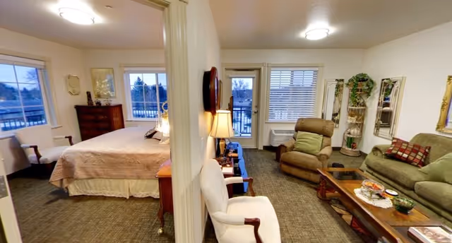 Interior view of a senior living facility suite divided into two sections by a partial wall. On the left side is a bedroom with a large bed, a wooden dresser, two windows, and a chair. On the right side is a living room area with a sofa, an armchair, a coffee table with decorative items, a lamp, a window, and a door leading outside. The room is carpeted and decorated with plants and mirrors on the walls.