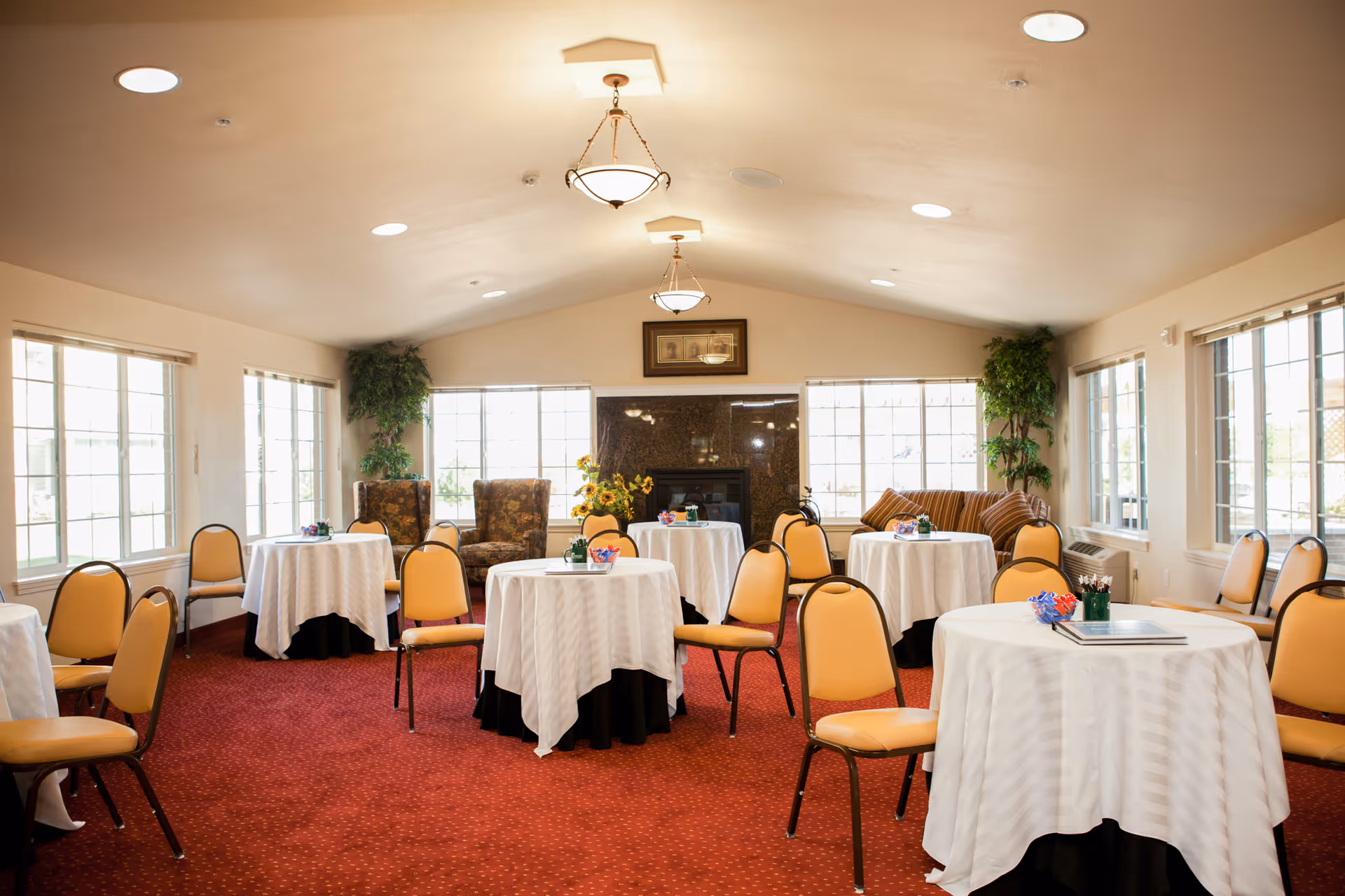 Bright community dining/activity room with round tables covered in white linens, yellow chairs, large windows, plants, and a fireplace.