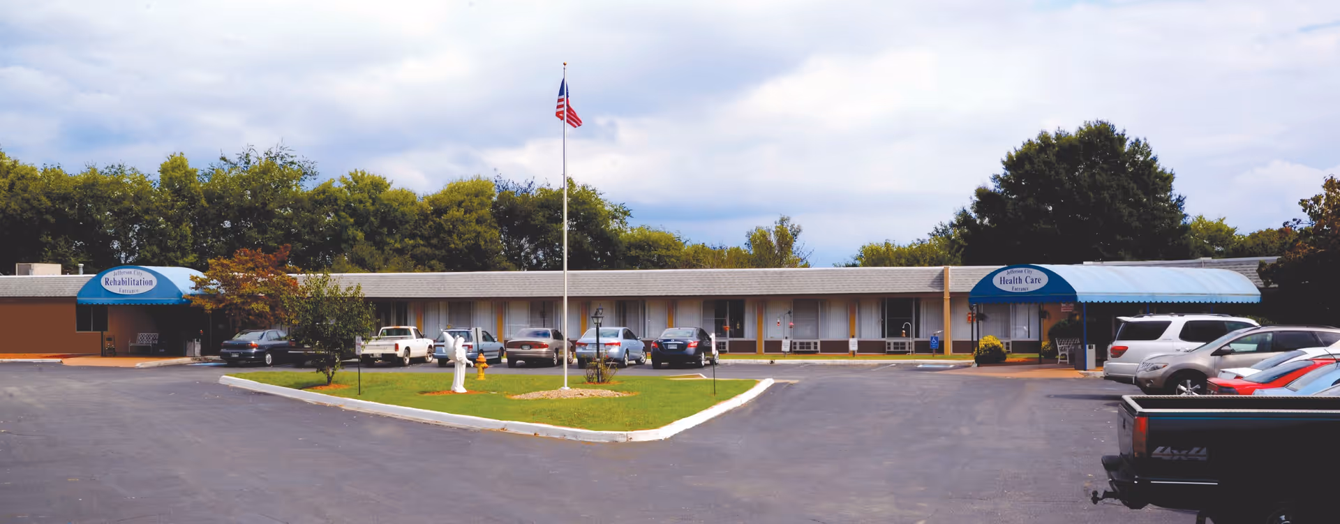 Exterior view of Jefferson City Health & Rehabilitation Center showing a single-story building with two blue awnings labeled Rehabilitation and Health Care. Several cars are parked in front of the building, and an American flag is flying on a flagpole in a grassy area with a statue and a fire hydrant. Trees and a cloudy sky are visible in the background.