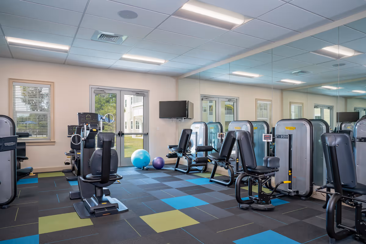 A fitness room with exercise machines including seated weight machines, two exercise balls on the floor, a wall-mounted TV, large windows, and a glass door leading outside. The room has a colorful tiled floor and a mirrored wall reflecting the equipment.