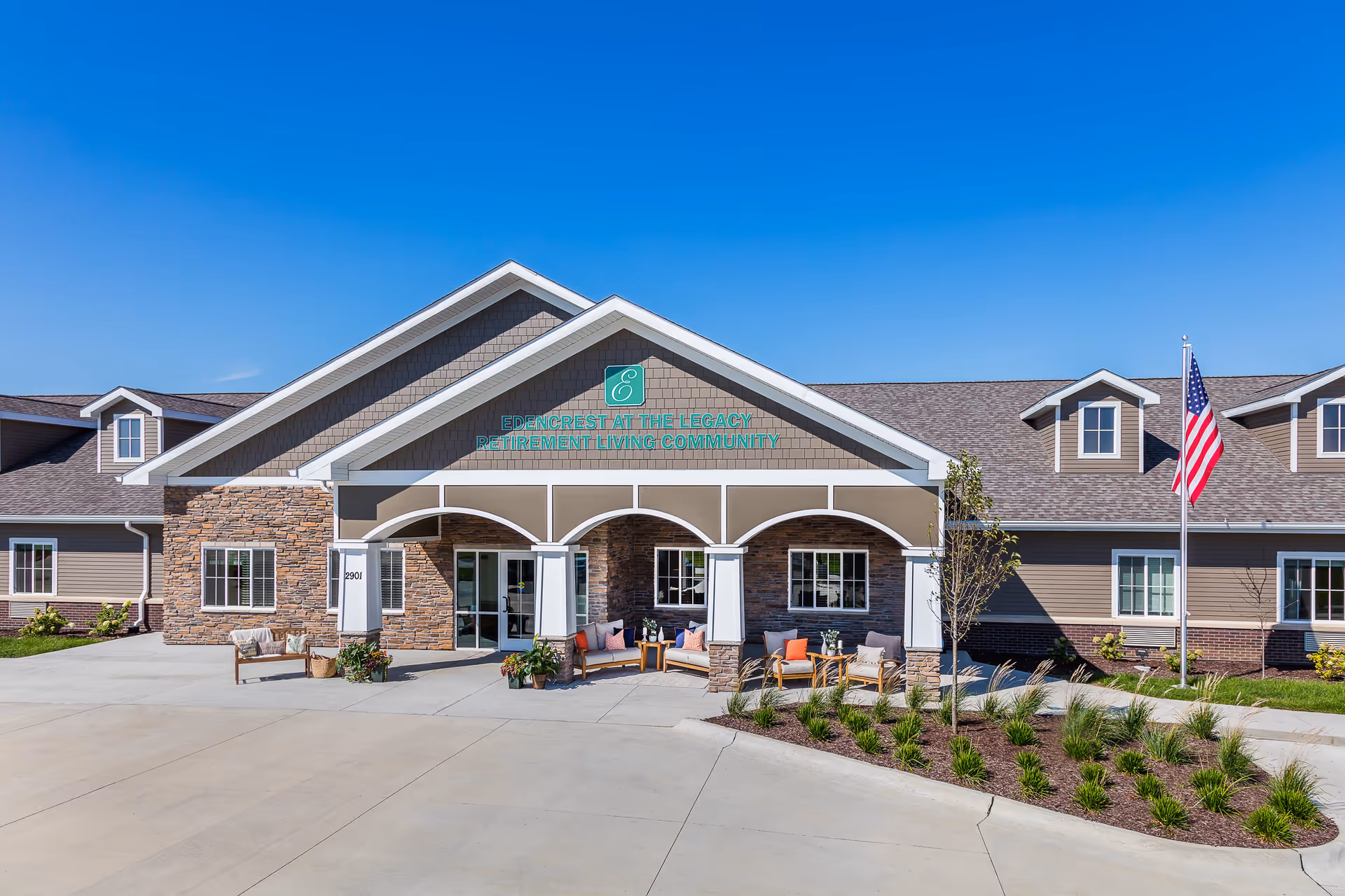 Front exterior view of Edencrest at The Legacy retirement living community building with a covered entrance, outdoor seating area with chairs and cushions, an American flag on a flagpole, and landscaping with grass and plants under a clear blue sky.