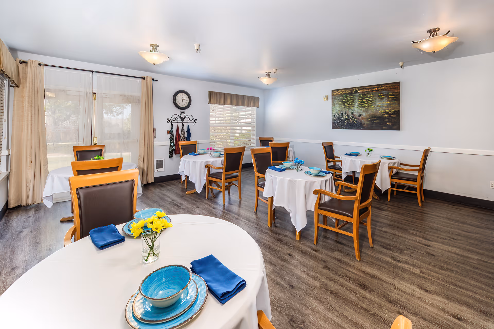 A bright dining room with several round tables covered with white tablecloths, each set with blue plates, bowls, and napkins. Wooden chairs with dark cushions surround the tables. The room has large windows with beige curtains, a wall clock, a coat rack with hanging items, and a decorative painting on the wall. The floor is a dark wood laminate.