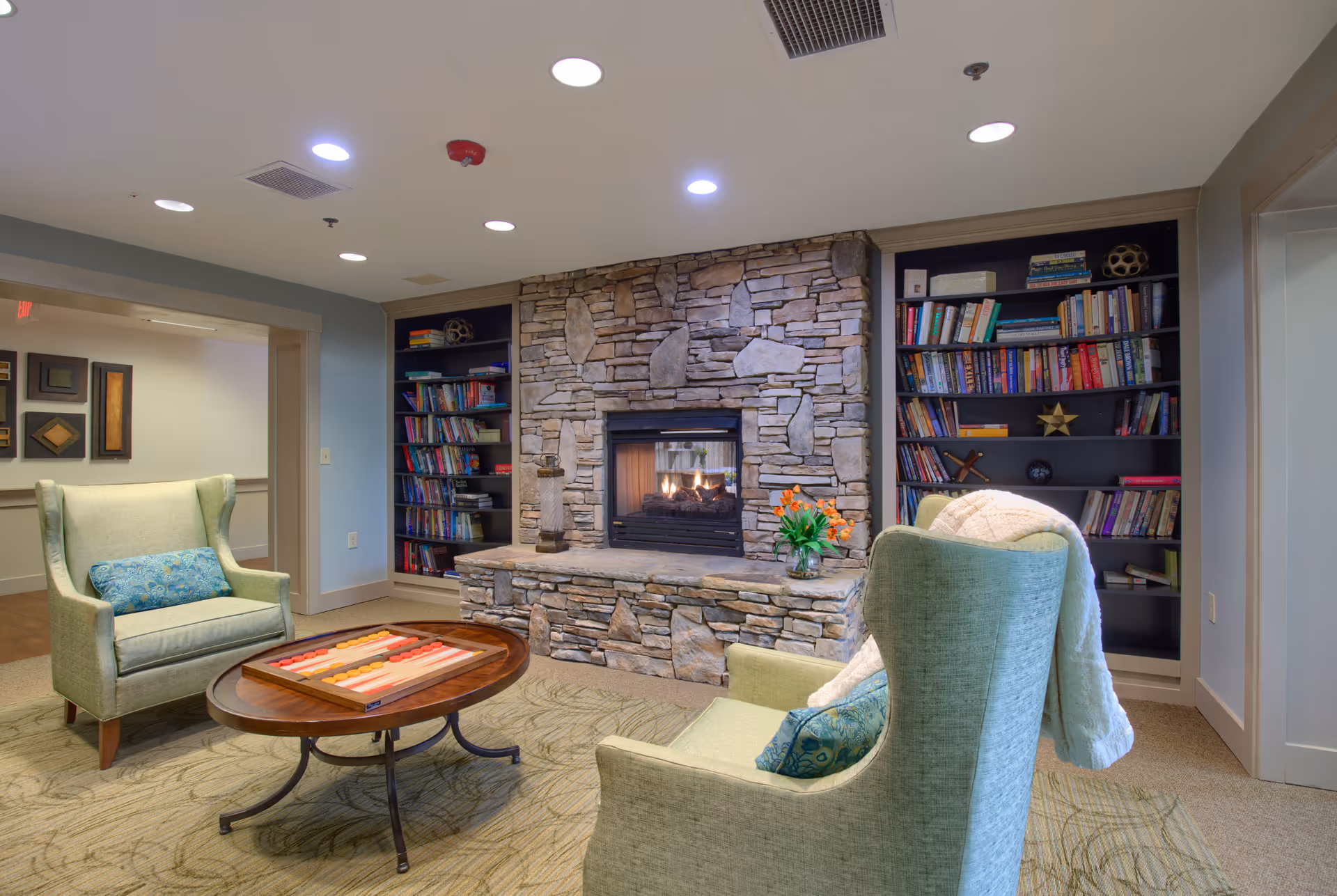 Cozy living room area with two green upholstered armchairs, a round wooden coffee table with a backgammon board, a stone fireplace with a fire burning, and built-in bookshelves filled with books on either side of the fireplace.