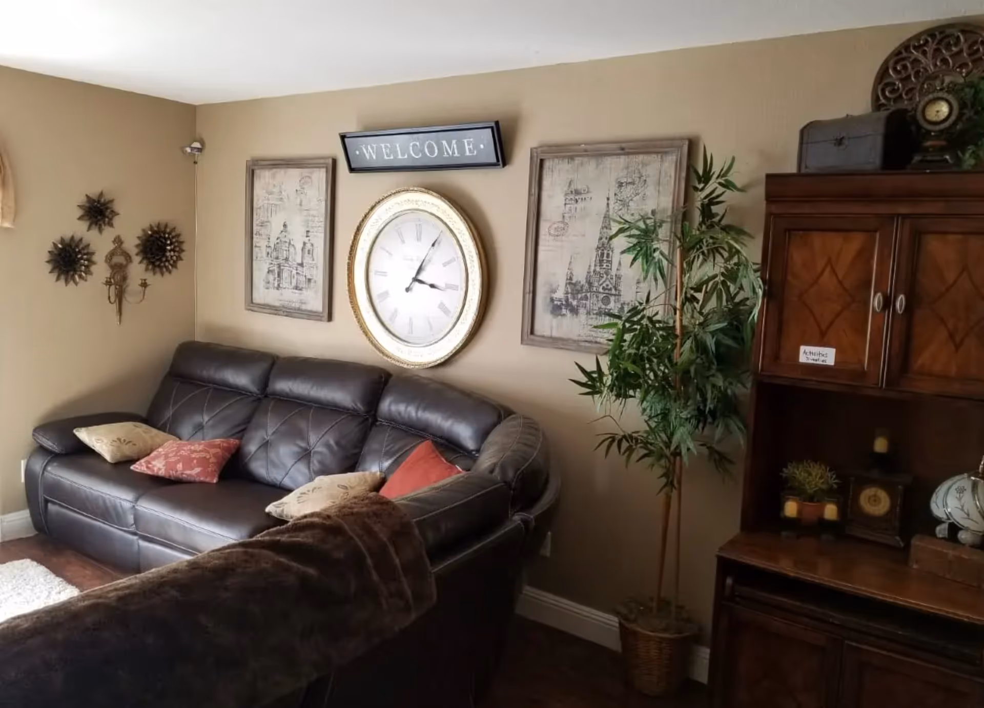 Cozy living room with a brown leather sofa, large wall clock and 'WELCOME' sign, decorative art, potted plant and wooden cabinet.
