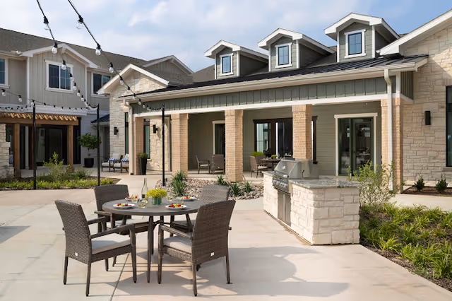 Sunlit outdoor courtyard with a round dining table and four wicker chairs, a built-in stone grill, string lights, and a covered patio of the building.
