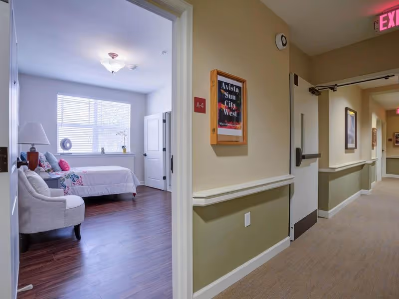 View down a hallway in a senior living facility with beige walls and carpeted floor. On the left, an open door reveals a bedroom with a bed, a chair, a lamp, and a window with blinds letting in natural light. A framed sign on the wall outside the bedroom reads 'Avista Sun City West'.