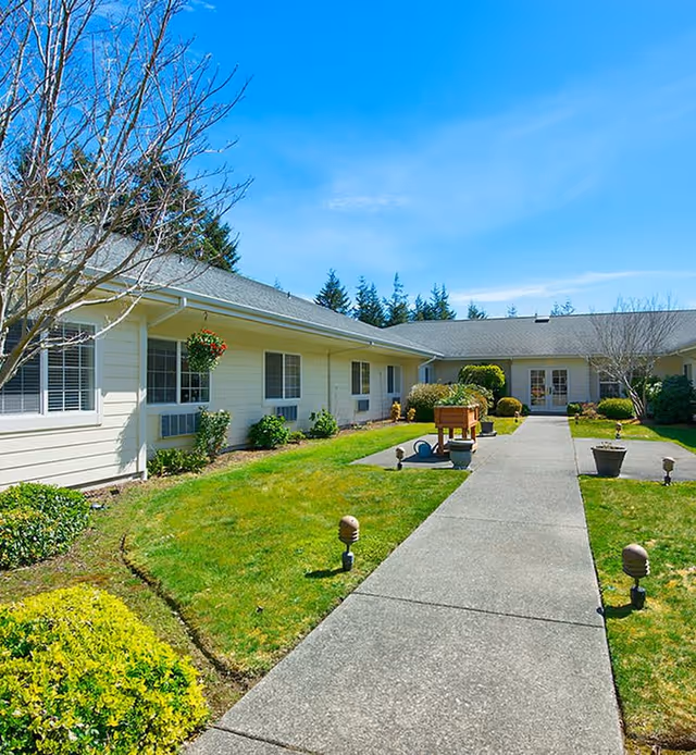 Sunny courtyard with a central concrete walkway flanked by grass, planters, and a one-story retirement community building.