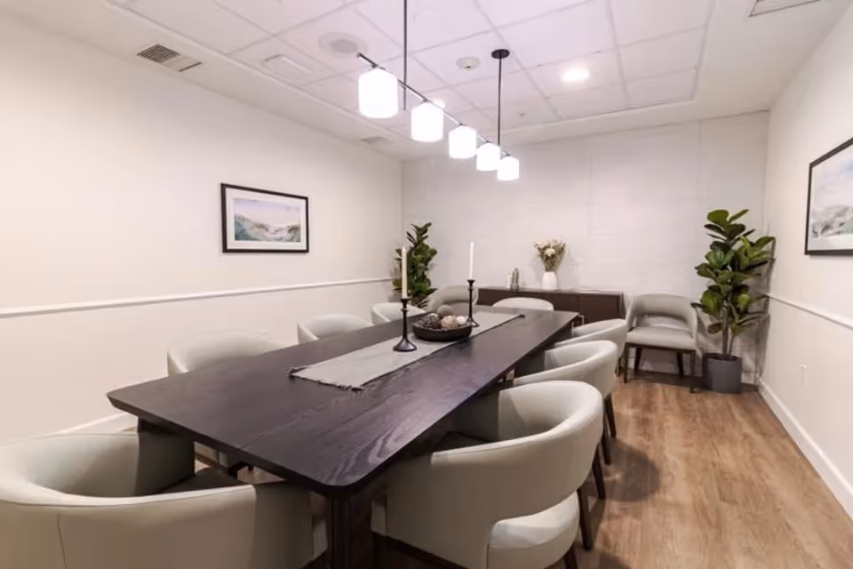 A modern dining room with a long dark wooden table surrounded by eight light gray upholstered chairs. Above the table hangs a row of five pendant lights with white shades. The room has light-colored walls with two framed landscape paintings and two potted plants in the corners. A sideboard with decorative items is against the far wall, and the floor is a light wood finish.