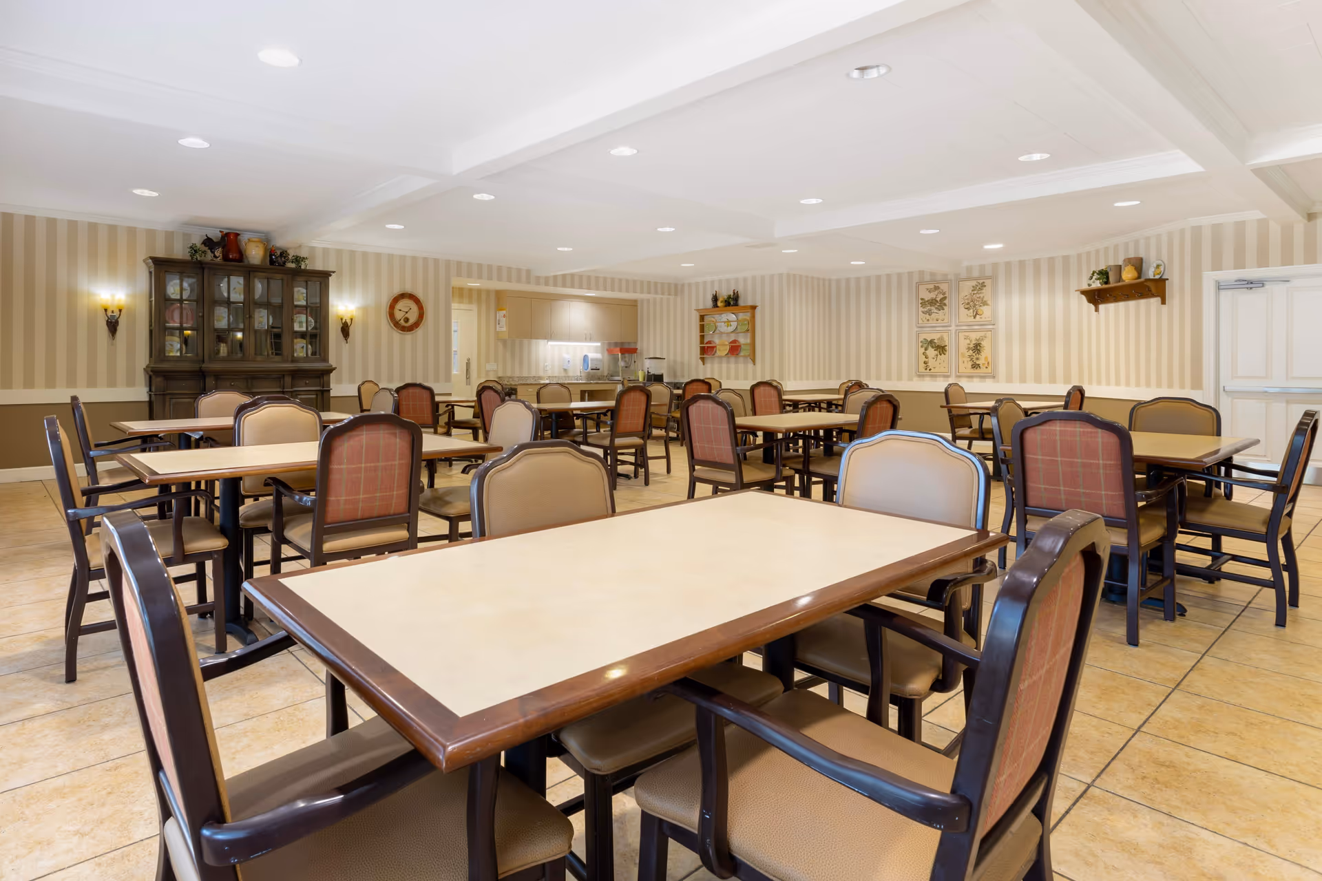 A spacious dining room with multiple tables and chairs arranged neatly. The room has beige striped wallpaper, tiled flooring, and soft lighting from ceiling fixtures and wall sconces. There is a wooden cabinet with glass doors on the left wall and framed botanical prints on the far wall. The overall atmosphere is warm and inviting.