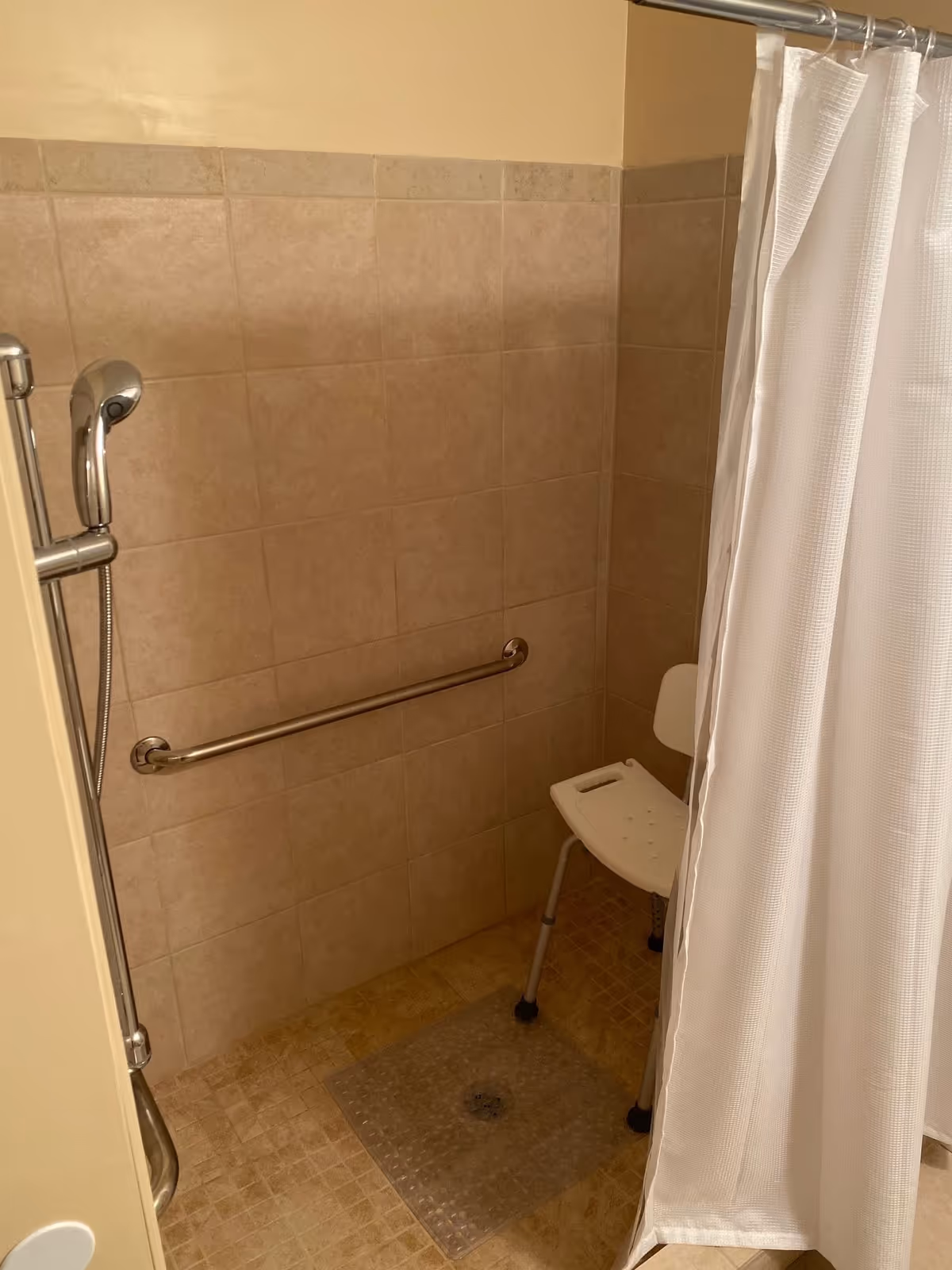 Tiled walk-in shower with a grab bar, handheld showerhead, a white shower curtain, and a shower chair.