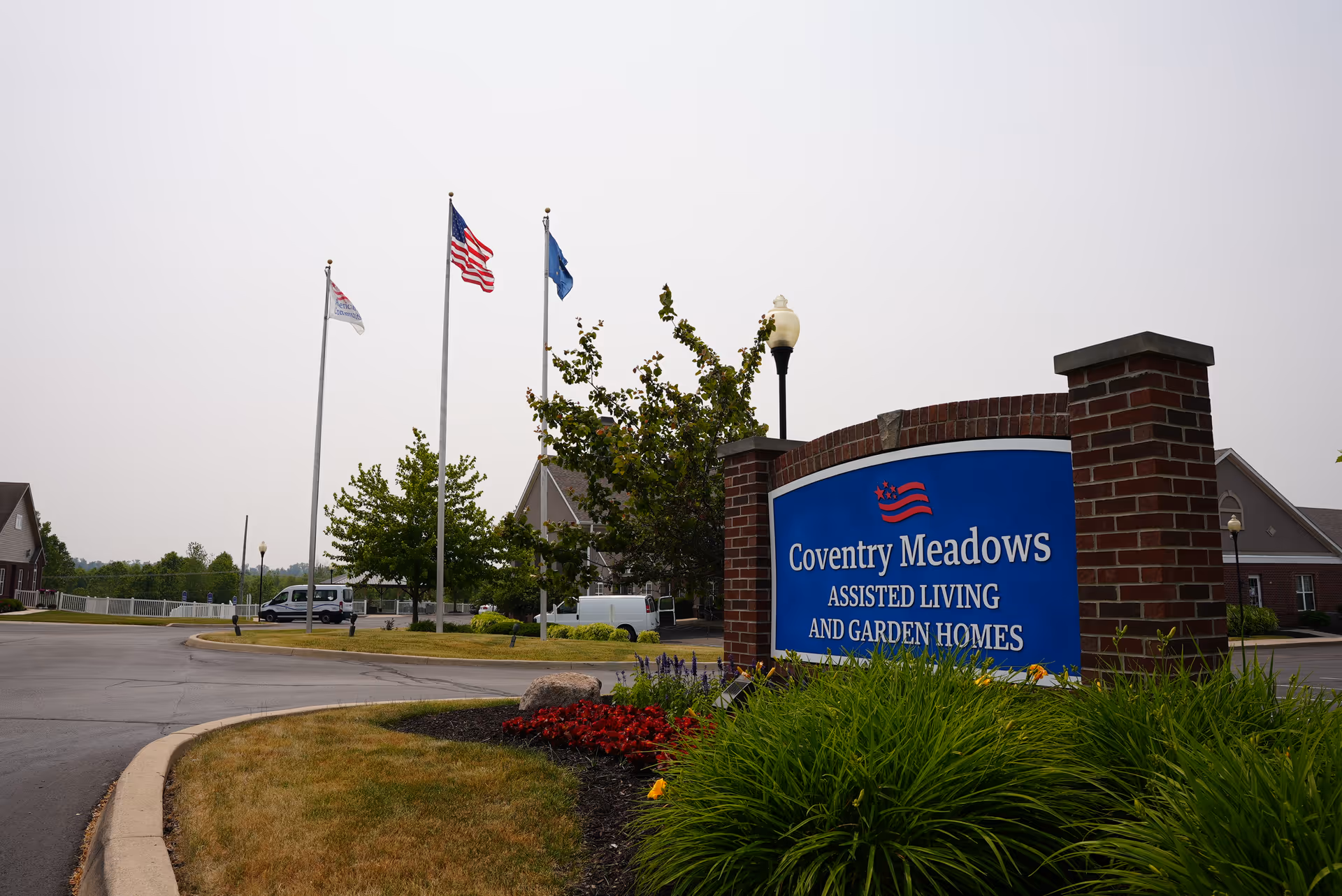 Entry sign for Coventry Meadows Assisted Living and Garden Homes at a landscaped roundabout with flagpoles and facility buildings in the background.