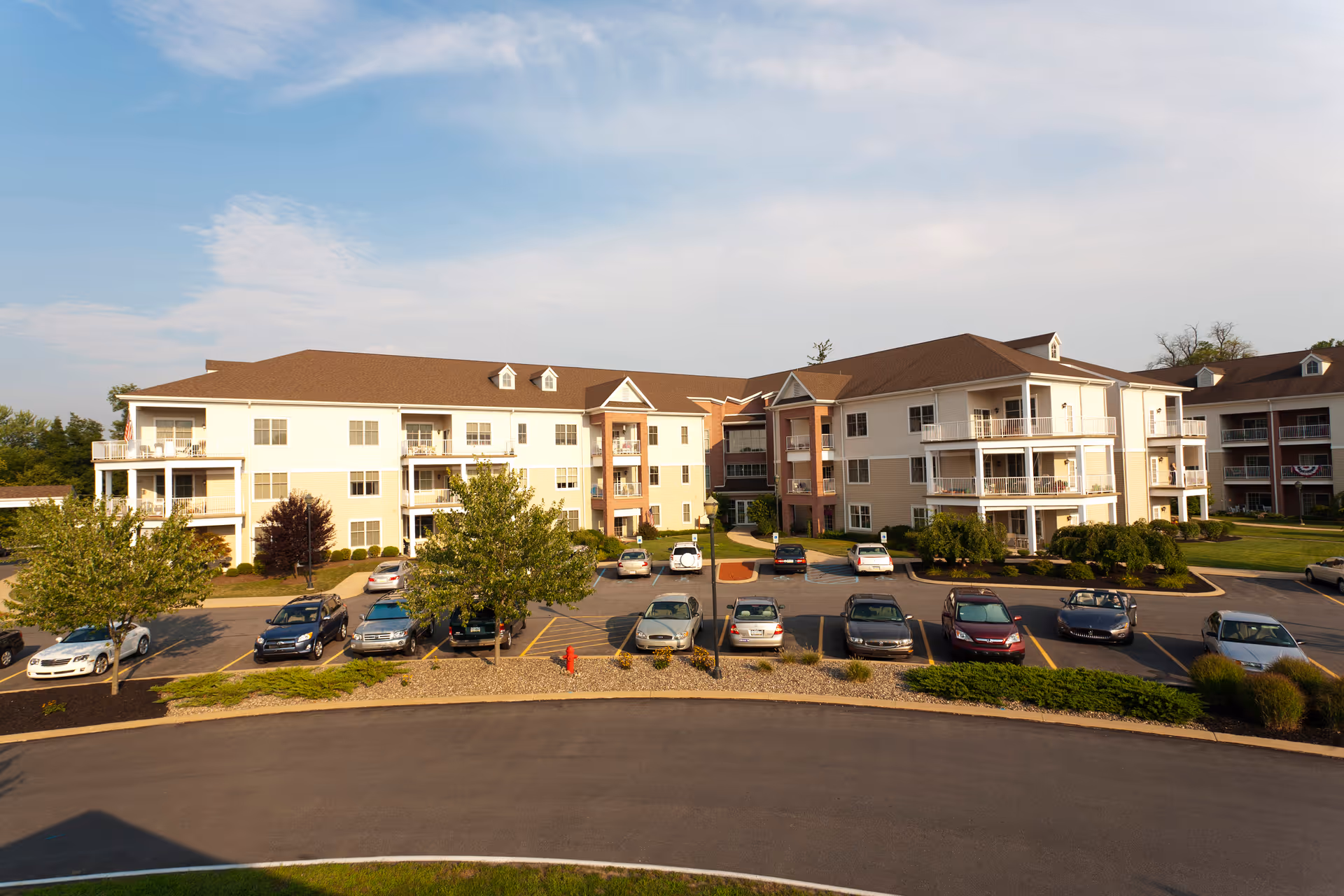 Exterior view of a three-story senior living community building with balconies, surrounded by a parking lot with several parked cars and landscaped greenery under a partly cloudy sky.