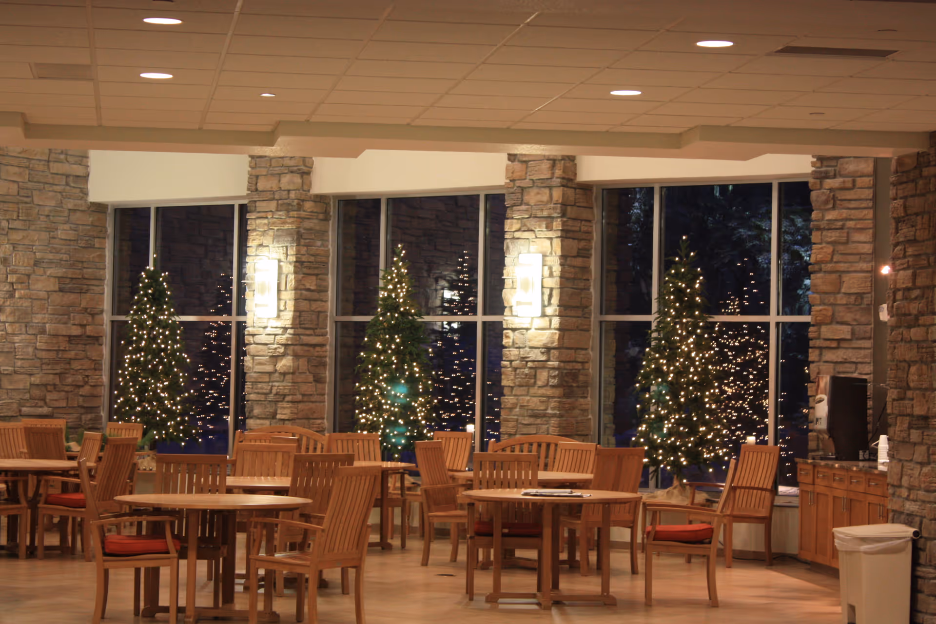 A spacious dining area with wooden tables and chairs featuring red cushions. The room has large windows with stone pillars between them, and several small Christmas trees decorated with white lights are placed in front of the windows. The ceiling has recessed lighting, and there is a counter with a coffee machine and a trash bin on the right side.
