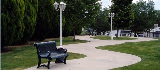 A winding concrete pathway in a green outdoor area with grass and trees on either side. There are black metal benches and white lamp posts along the path.