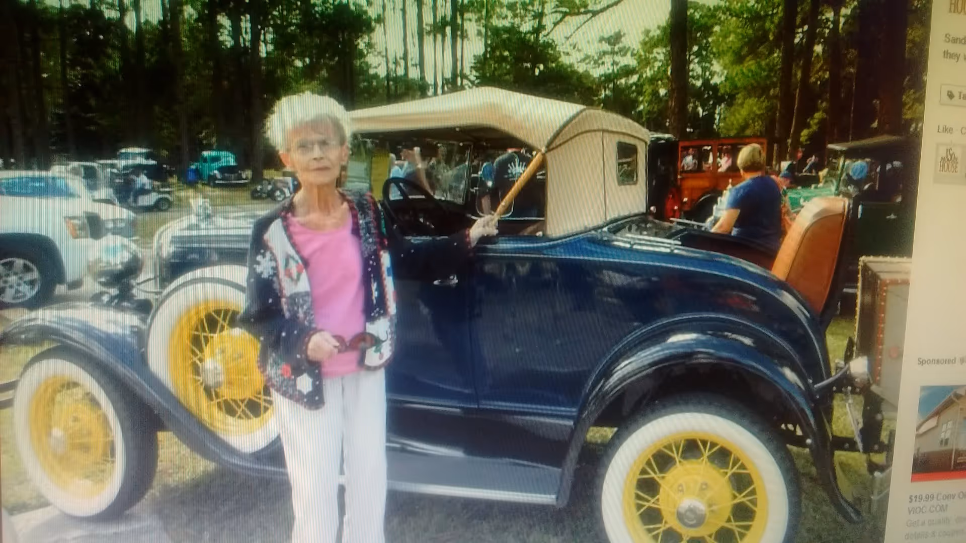 An elderly woman with white hair wearing a pink shirt, white pants, and a patterned cardigan stands next to a vintage dark blue car with yellow wheels and a beige convertible top. The background shows several other vintage cars and people in an outdoor setting with trees.