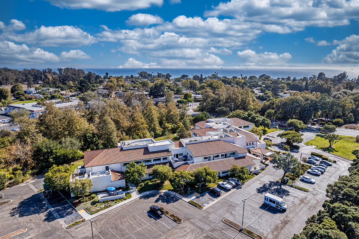 Aerial view of GranVida Senior Living and Memory Care facility showing a large building with red-tiled roofs surrounded by trees and greenery. There is a parking lot with several cars and a van parked. In the background, there are residential houses and a partly cloudy blue sky.