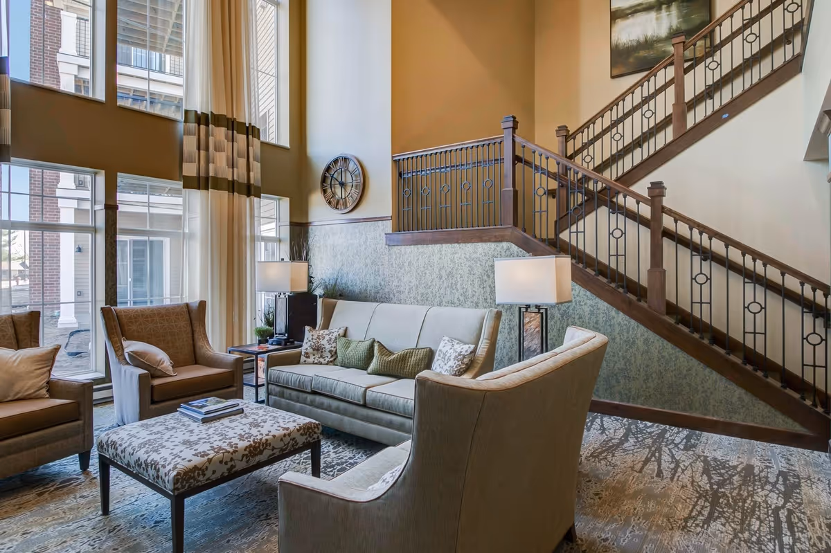A spacious and well-lit living room area with high ceilings and large windows. The room features a beige sofa with decorative pillows, two armchairs, a patterned ottoman, two table lamps, and a wooden staircase with metal railings leading to an upper floor. A large wall clock and a framed painting are visible on the walls.