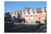 Exterior view of a multi-story senior living facility building with a covered entrance, several windows, and parked cars in front under a clear blue sky.
