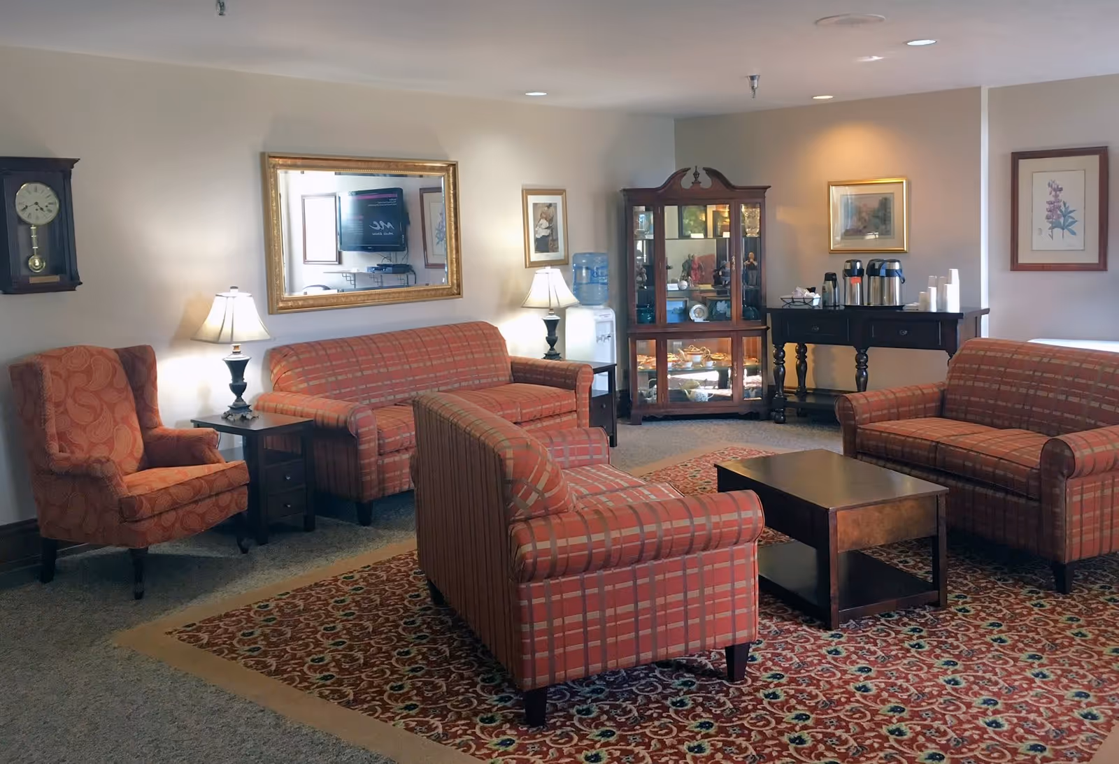 A cozy living room with patterned red sofas and an armchair arranged around a wooden coffee table on a decorative rug. The room features a wall clock, two table lamps, framed artwork, a large mirror, a glass display cabinet with decorative items, a water dispenser, and a table with coffee urns and cups.