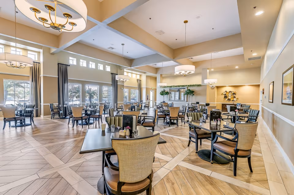 A spacious and well-lit dining room with multiple tables and chairs arranged neatly. The room features large windows with curtains, modern chandeliers hanging from the ceiling, and decorative plants and artwork on the walls. The floor has a patterned wood design.