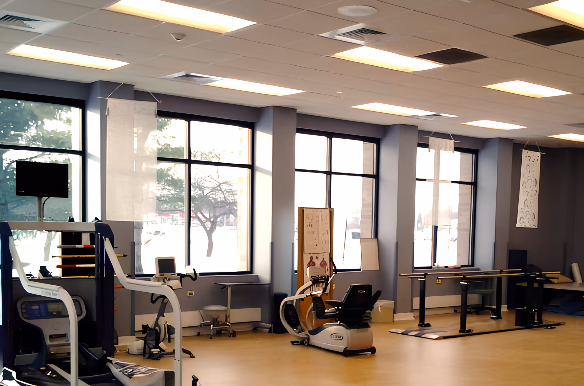 A spacious rehabilitation room with large windows letting in natural light. The room contains exercise equipment including a recumbent bike and parallel bars for physical therapy. Anatomical charts are displayed on the walls.