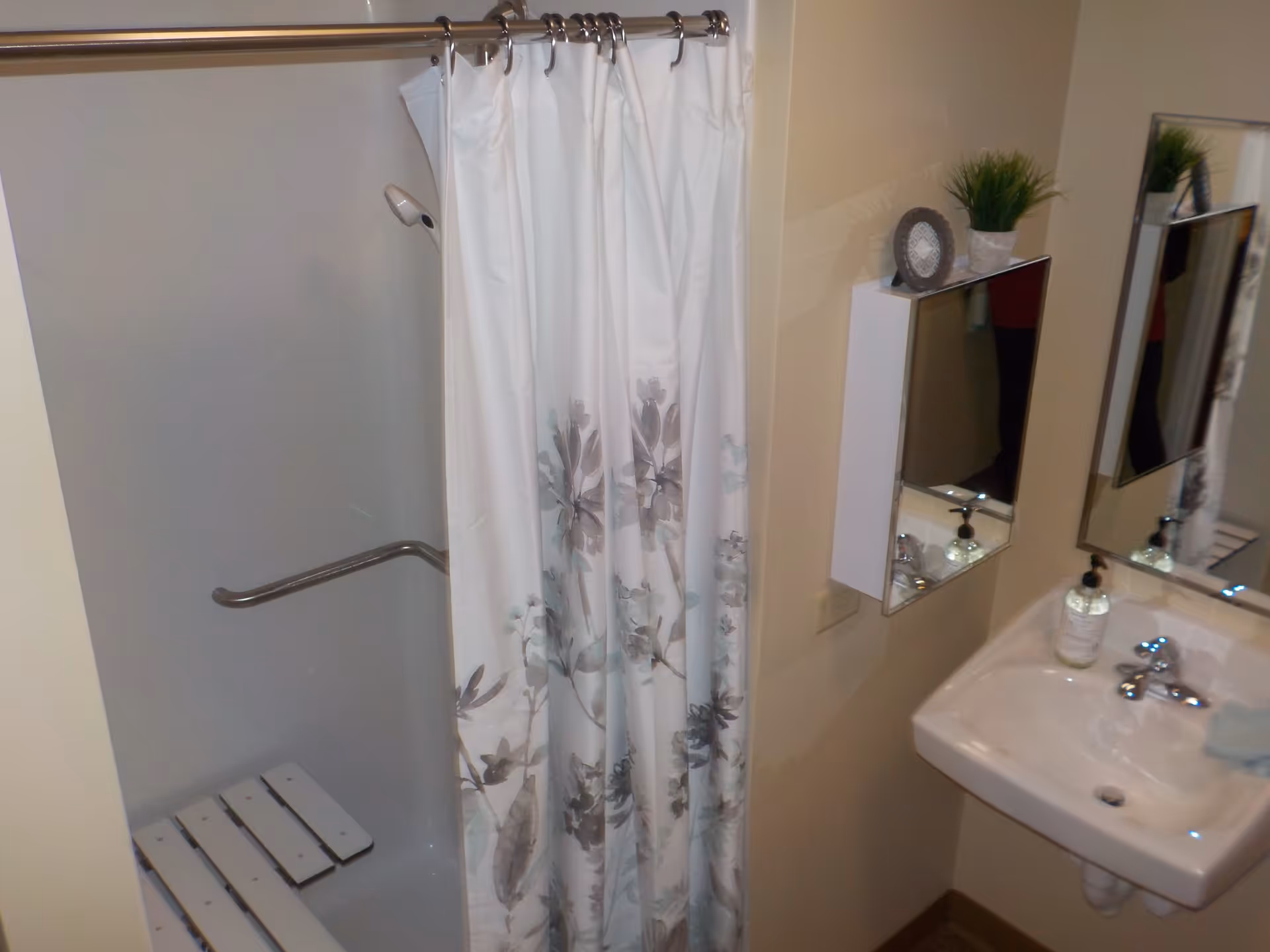 A bathroom with a shower area featuring a white shower curtain with floral patterns, a wall-mounted grab bar, and a foldable shower seat. Next to the shower is a white sink with a soap dispenser, a mirror above it, and a small wall-mounted cabinet with a decorative plant and a framed item on top.