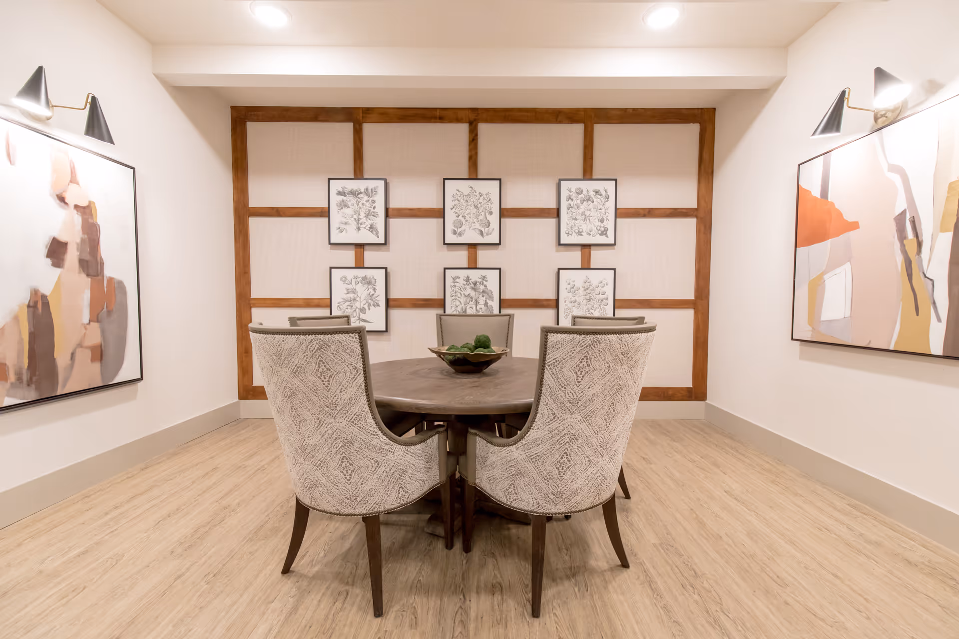 A modern dining room with a round wooden table surrounded by six upholstered chairs. The walls are decorated with framed botanical prints and abstract art paintings, and the floor has light wood flooring.