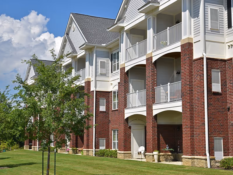 Exterior view of a multi-story assisted living facility building with red brick and white siding, featuring balconies and a well-maintained lawn with trees under a blue sky with clouds.