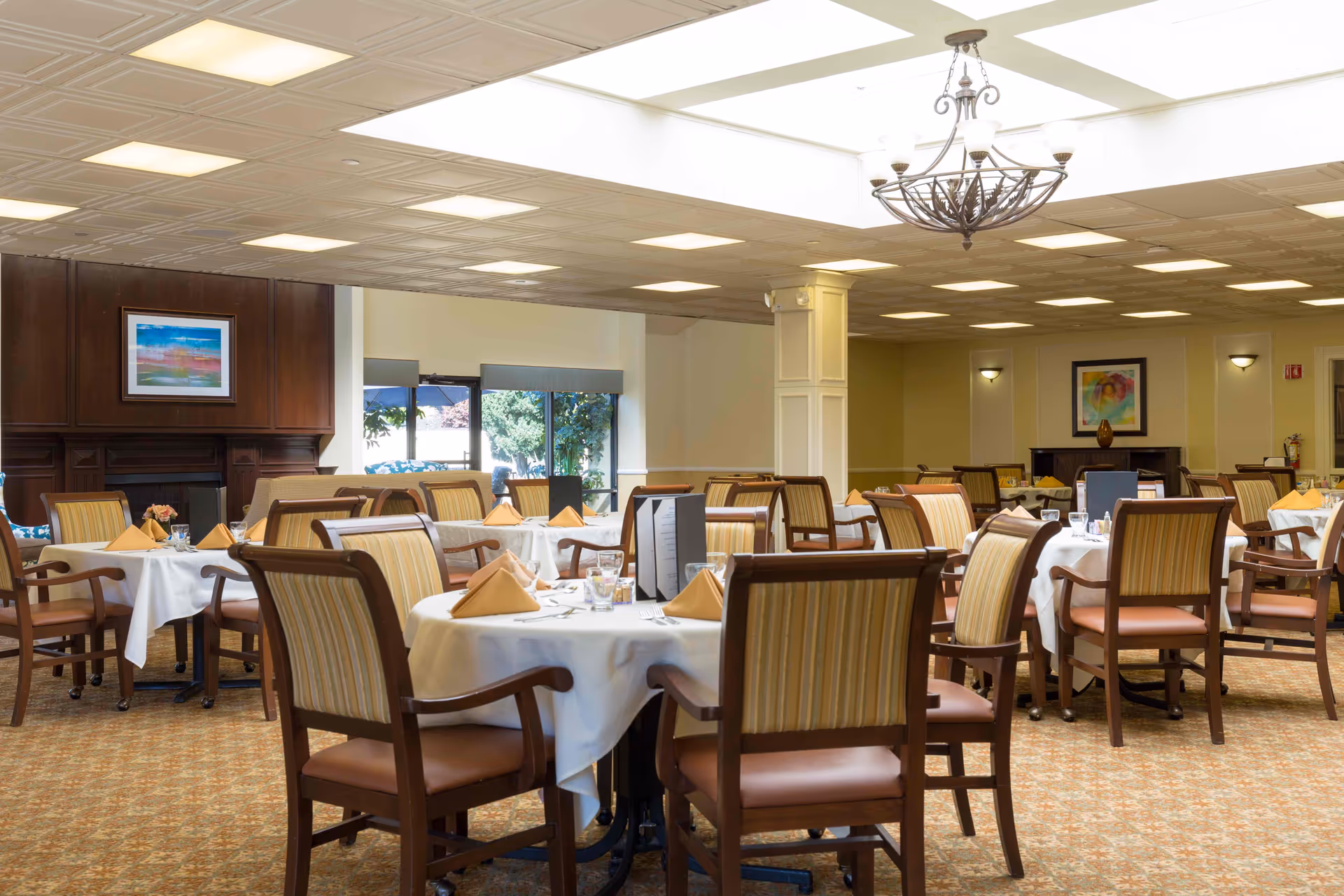 A bright dining room with multiple round tables set with white tablecloths and folded napkins, surrounded by wooden chairs and a chandelier overhead.