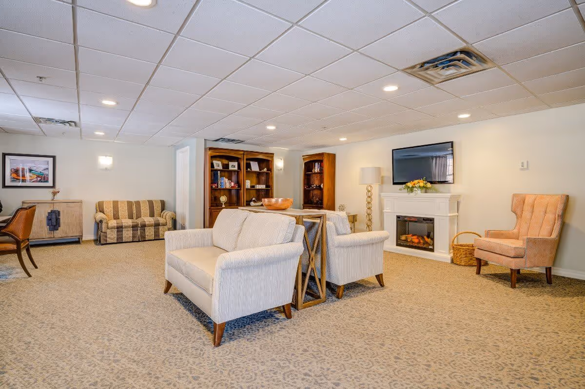 A spacious living room area with beige carpet and white walls. The room features two white armchairs and a white loveseat arranged around a small wooden table. In the background, there is a striped sofa, wooden bookshelves, a white electric fireplace with a flat-screen TV mounted above it, a tall floor lamp, and a peach-colored armchair with a basket beside it. The ceiling has recessed lighting and white tiles.