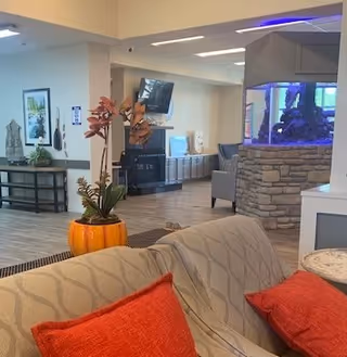 Interior common lounge with sofas and orange pillows, a potted plant, a wall-mounted TV, and a large aquarium set in a stone pillar.