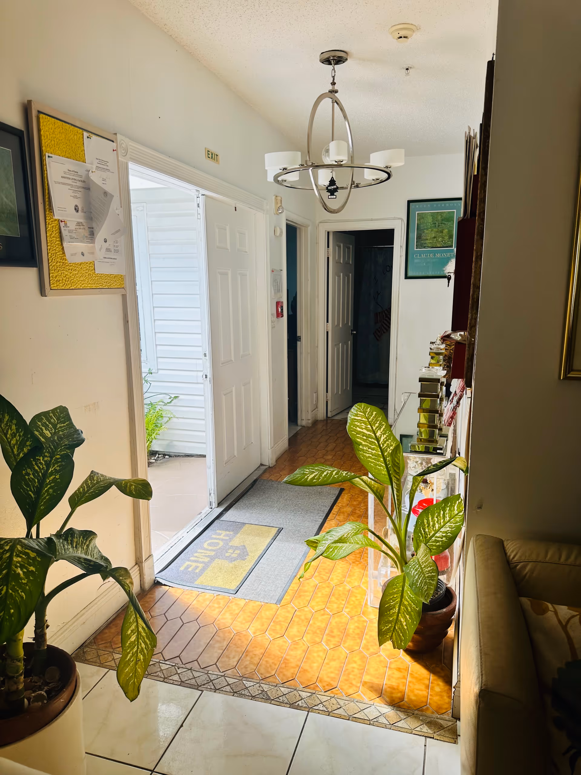 Interior hallway of a senior living facility with a tiled floor and a gray mat that says 'HOME' near an open door leading outside. There are two large potted green plants on either side of the hallway, a bulletin board with papers on the left wall, a modern chandelier hanging from the ceiling, and a framed picture on the far wall. A beige couch is partially visible on the right side.