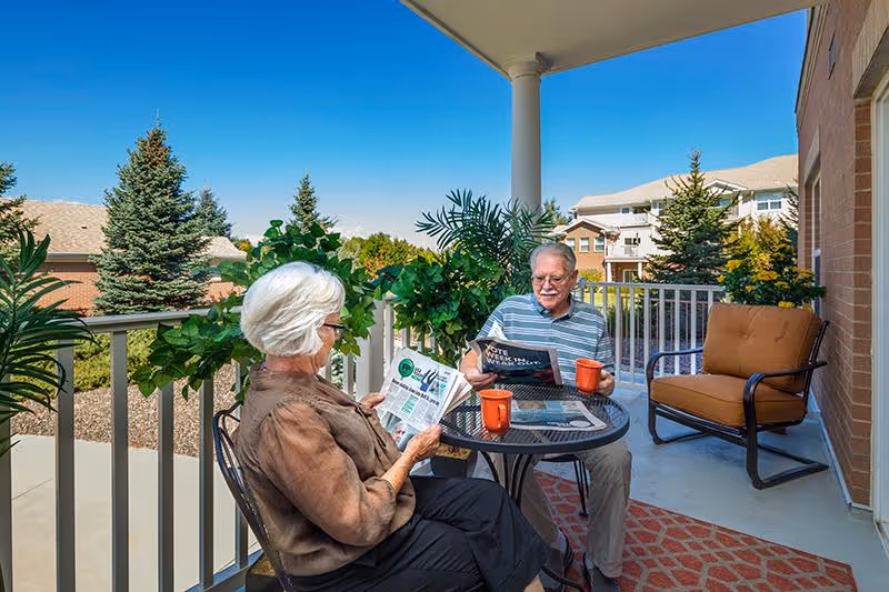An elderly man and woman sitting on a covered outdoor patio at a round glass table, reading newspapers and drinking from orange mugs. The patio has a railing with green plants and overlooks a landscaped area with trees and neighboring buildings under a clear blue sky.