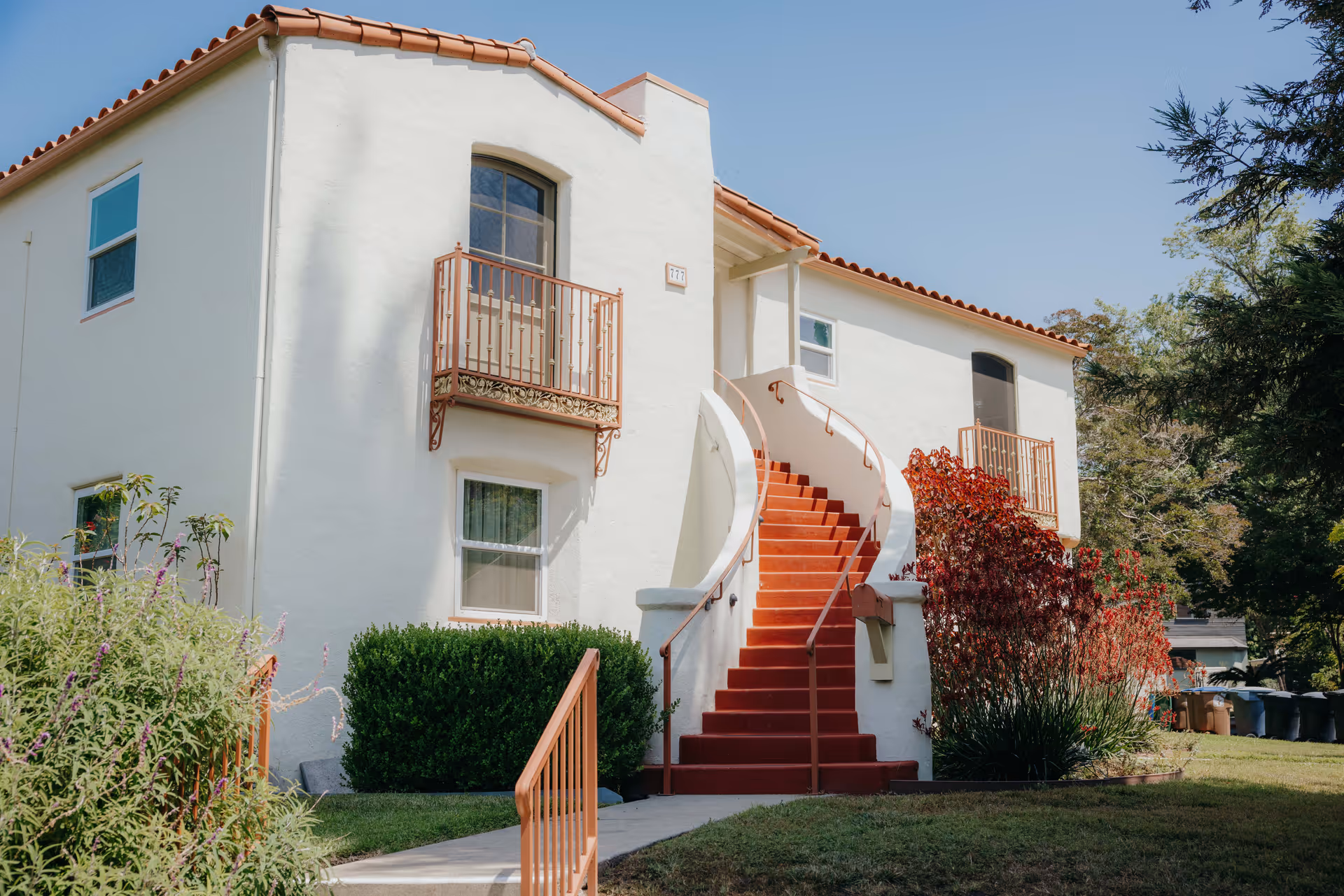 White two-story stucco building with a red tile roof and a curved red staircase leading to second-floor entrances, flanked by small balconies and landscaping.