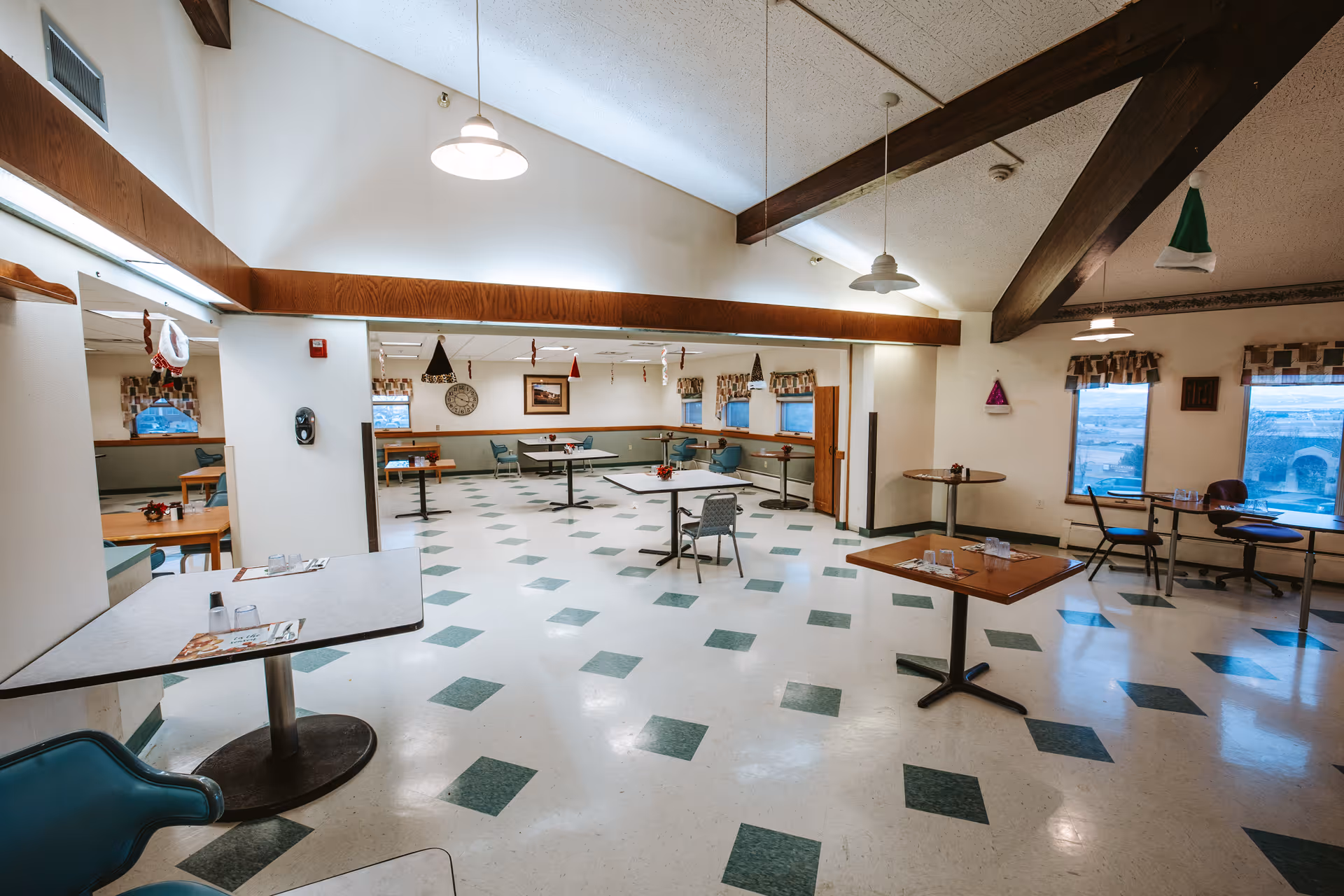 A spacious dining room with multiple tables and chairs arranged throughout. The floor has a white and green checkered pattern. The room features high ceilings with exposed wooden beams and hanging pendant lights. Windows with patterned valances allow natural light to enter. Some holiday decorations hang from the ceiling beams.