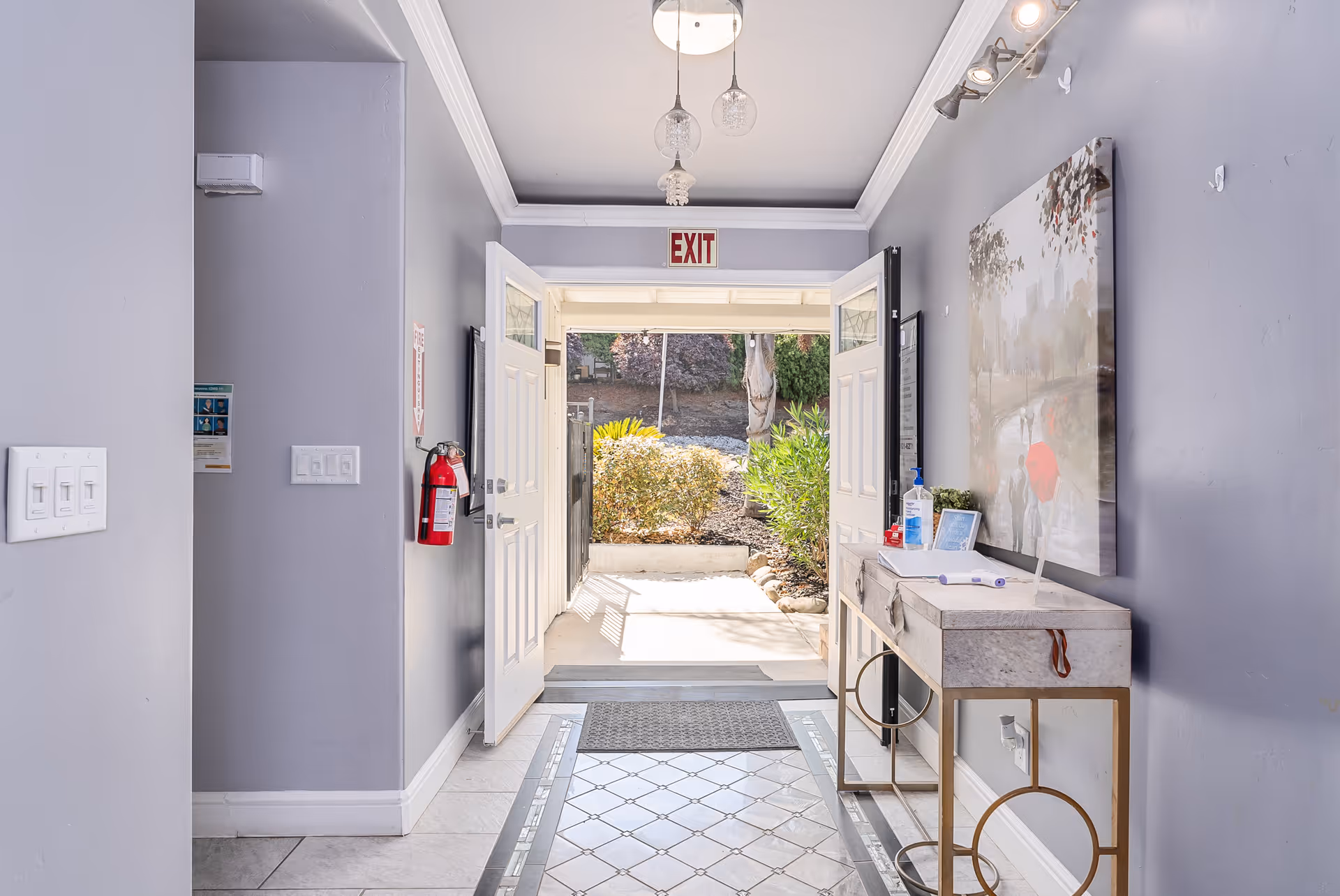View of an entrance hallway with double doors open to the outside. The hallway has light gray walls, a tiled floor with a decorative pattern, a small console table with hand sanitizer and a framed picture, a fire extinguisher mounted on the left wall, and an exit sign above the doors. Outside, there are bushes and trees visible.