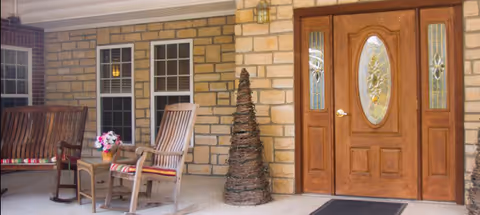A covered outdoor porch area with two wooden rocking chairs with striped cushions, a small wooden table with a flower arrangement, a tall woven decorative piece, and a wooden front door with decorative glass panels. The wall is made of light-colored stone with two windows.
