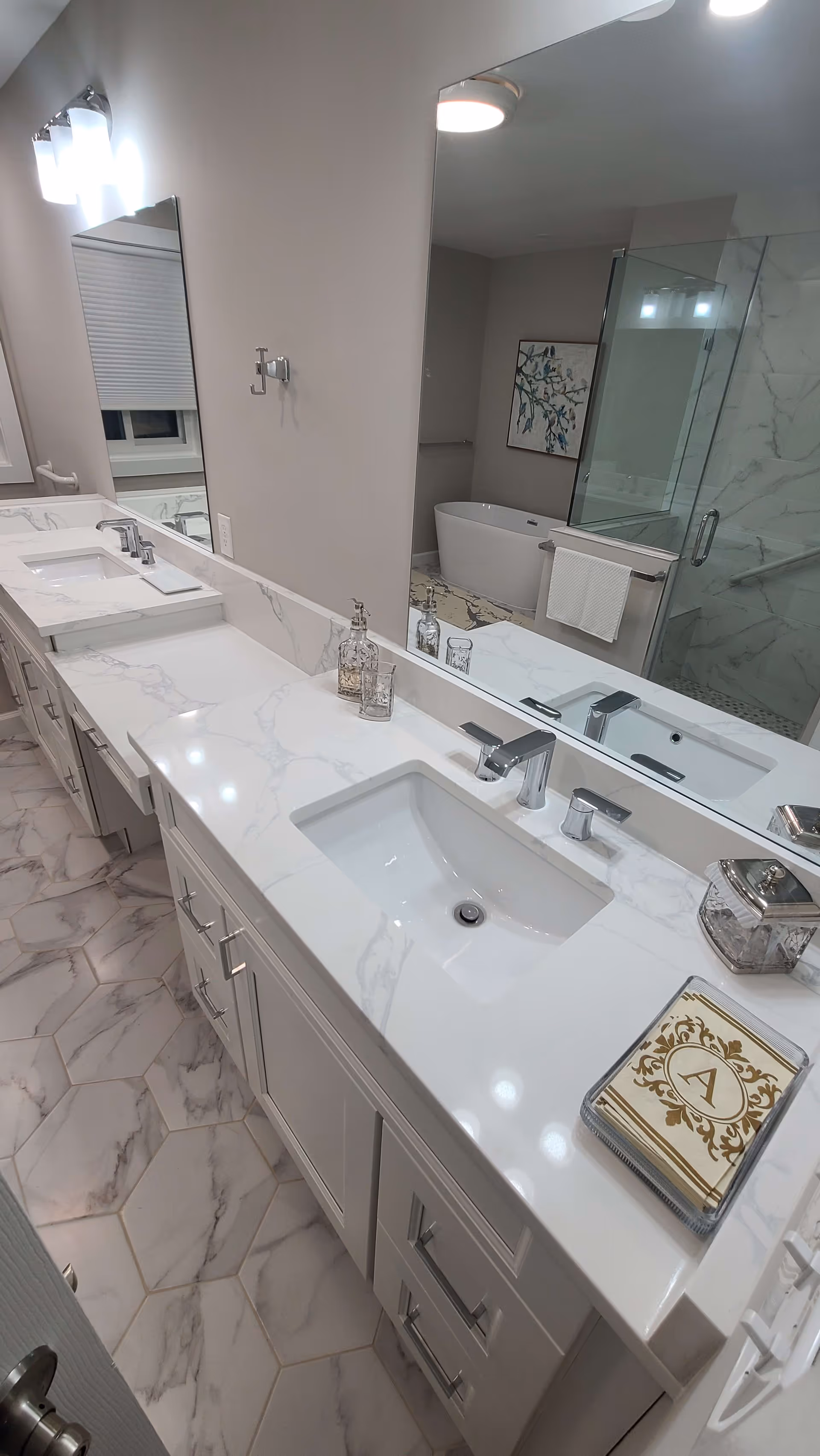 A modern bathroom with a long white marble countertop featuring two sinks and chrome faucets. The floor has hexagonal marble tiles. A large mirror runs along the wall above the countertop. In the background, there is a glass-enclosed shower and a freestanding bathtub with a towel hanging on a rack. A decorative framed artwork is visible on the wall near the bathtub.