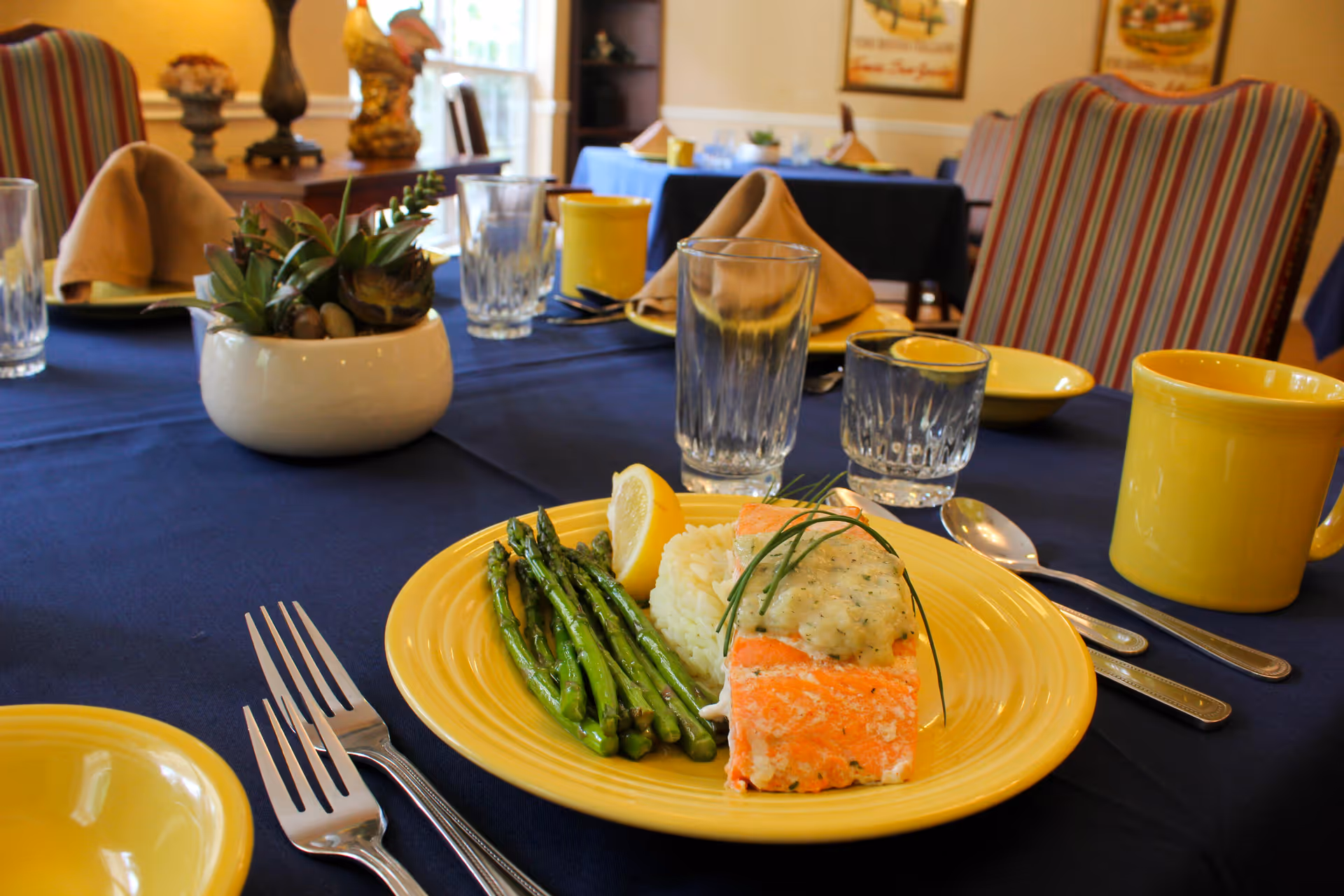 A dining table set with a yellow plate containing cooked salmon topped with a creamy sauce, grilled asparagus, a lemon wedge, and a serving of rice. The table is covered with a dark blue tablecloth and has yellow mugs, clear drinking glasses, silverware, and a small white pot with green succulent plants. In the background, there are striped upholstered chairs and framed pictures on the wall.