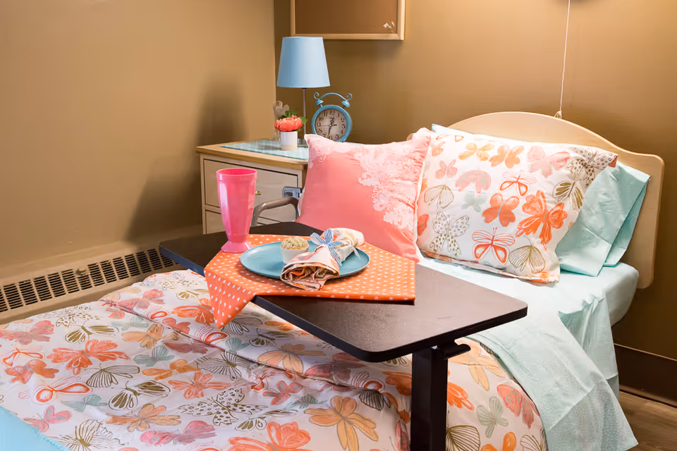 A hospital-style bed with butterfly-patterned bedding and colorful pillows, a bedside table with a blue lamp and an alarm clock, and a tray table holding a pink cup, a plate with a napkin, and a muffin.