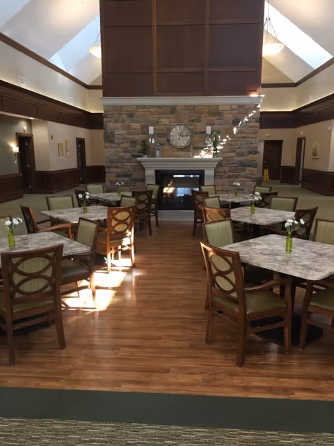 A dining room with multiple square tables and wooden chairs with green cushions. Each table has a small vase with white flowers. The room features a stone fireplace with a clock above it, wood-paneled walls, and a high ceiling with skylights allowing natural light to enter.