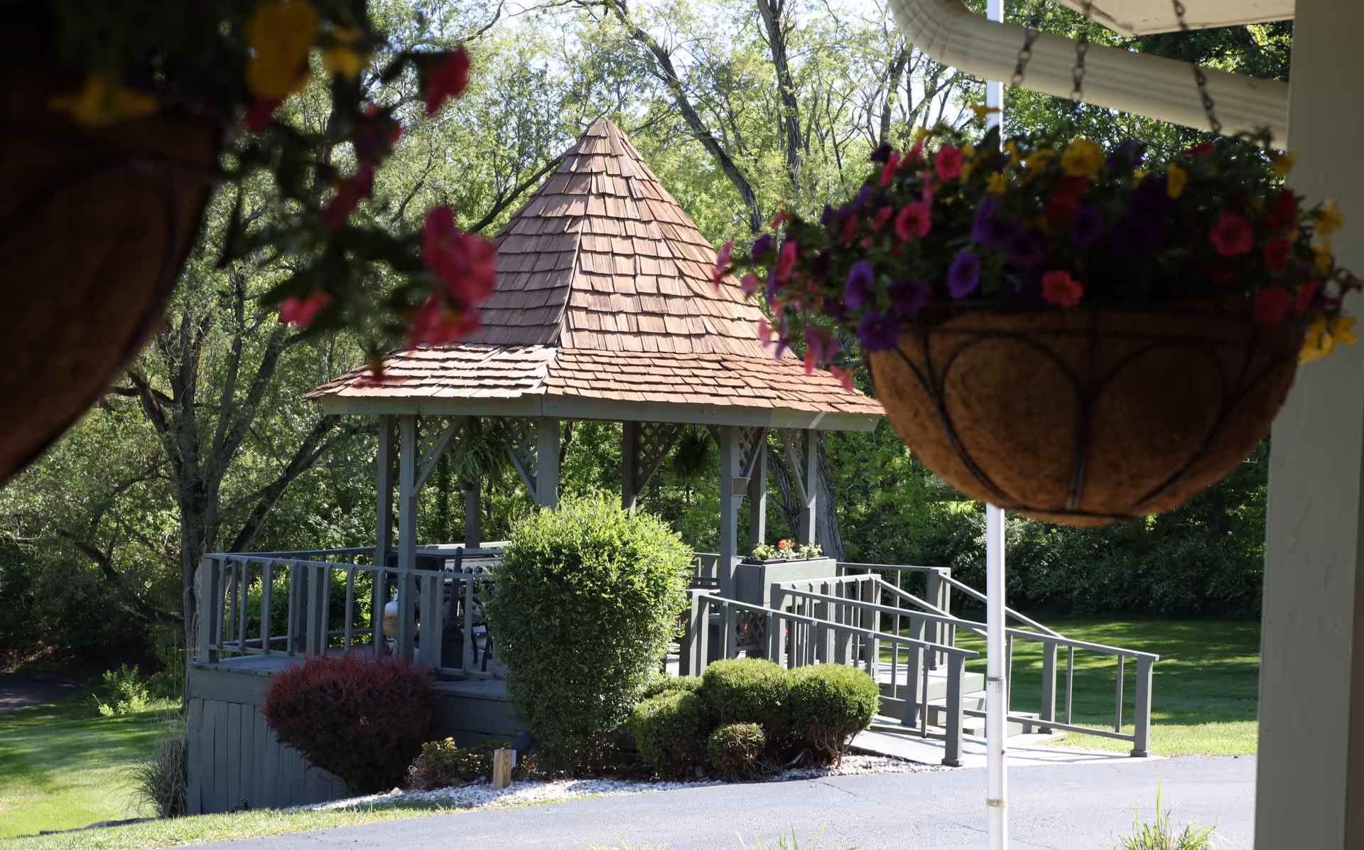 A wooden gazebo with a shingled roof surrounded by green bushes and trees in a garden setting. Hanging flower baskets with colorful flowers are visible in the foreground.