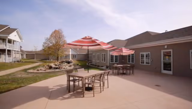 Outdoor patio area at Trail Ridge Senior Living Community with tables and chairs under red and white striped umbrellas, surrounded by buildings and a small landscaped area with a tree and rocks.