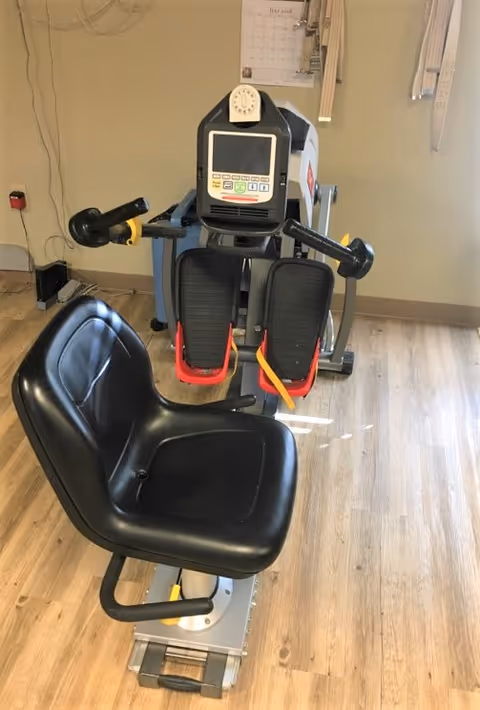 A black exercise machine with a seat and foot pedals in a room with light wood flooring and beige walls. The machine has a digital display and handles on either side. A calendar is visible on the wall behind the machine.