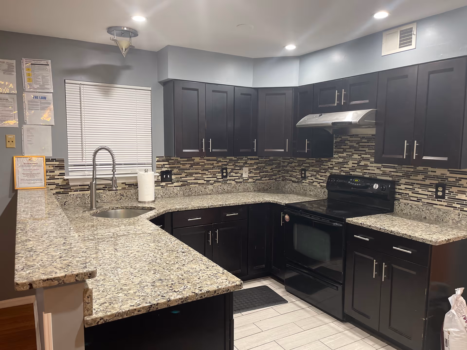 Modern kitchen with dark wood cabinets, granite countertops, a stainless steel sink with a high-arc faucet, a black electric stove with an overhead stainless steel range hood, and a mosaic tile backsplash in shades of black, gray, and beige. The kitchen has light-colored tiled flooring and gray walls with a window covered by white blinds.
