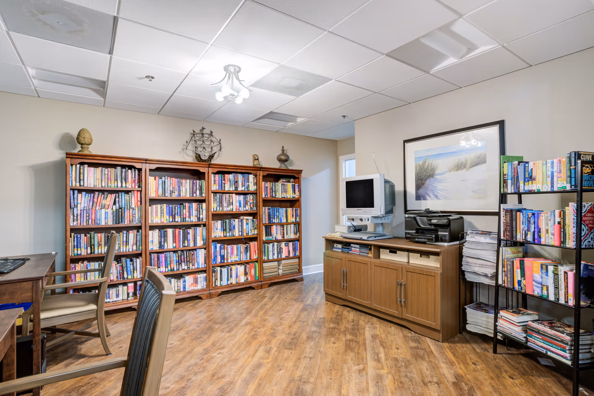 Interior room with wooden bookshelves filled with books, a wooden cabinet holding a computer monitor and printer, a metal bookshelf with more books and magazines, and several chairs around a wooden table. The room has wood flooring and a framed beach-themed picture on the wall.
